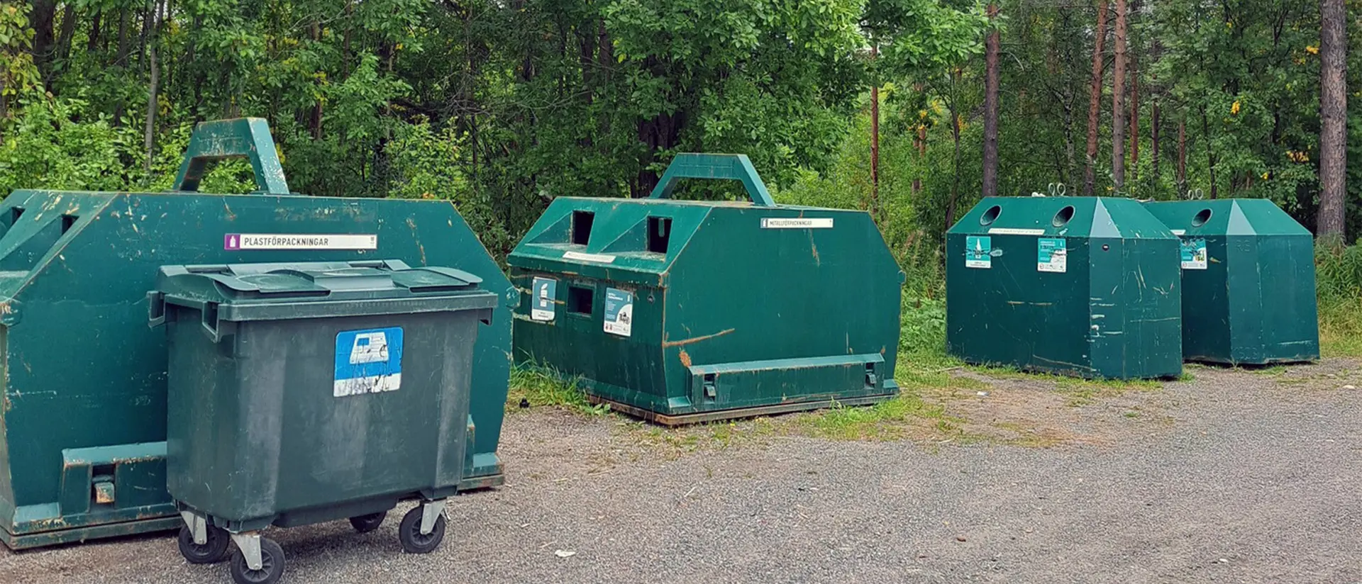 Återvinningsstation med en separat plasttunna för tidningar framför. Bilden är tagen på sommaren och träden är gröna. Foto. 