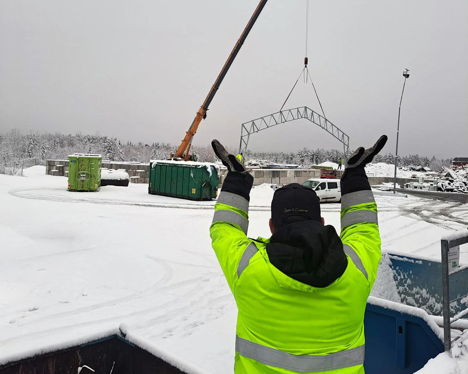 En person i varselkläder sett bakifrån håller upp händerna för att visualisera hur konstruktionen tar form. I bakgrunden syns bygget av tälthallen med en lyftkran och första stålkonstruktion på plats. Det är snö på backen. Foto