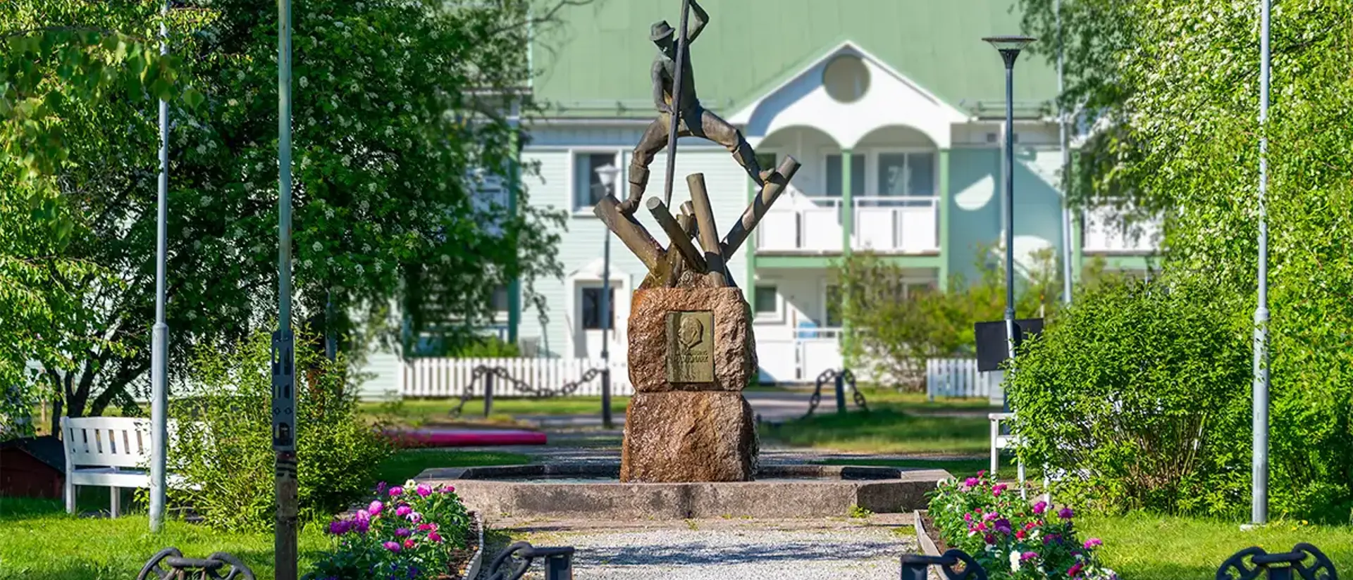 Skulpuren av en timmerflottare vid fontänen i parken i jokkmokk. Bilden är tagen en sommardag med blommor, grönska och blå himmel. Foto.