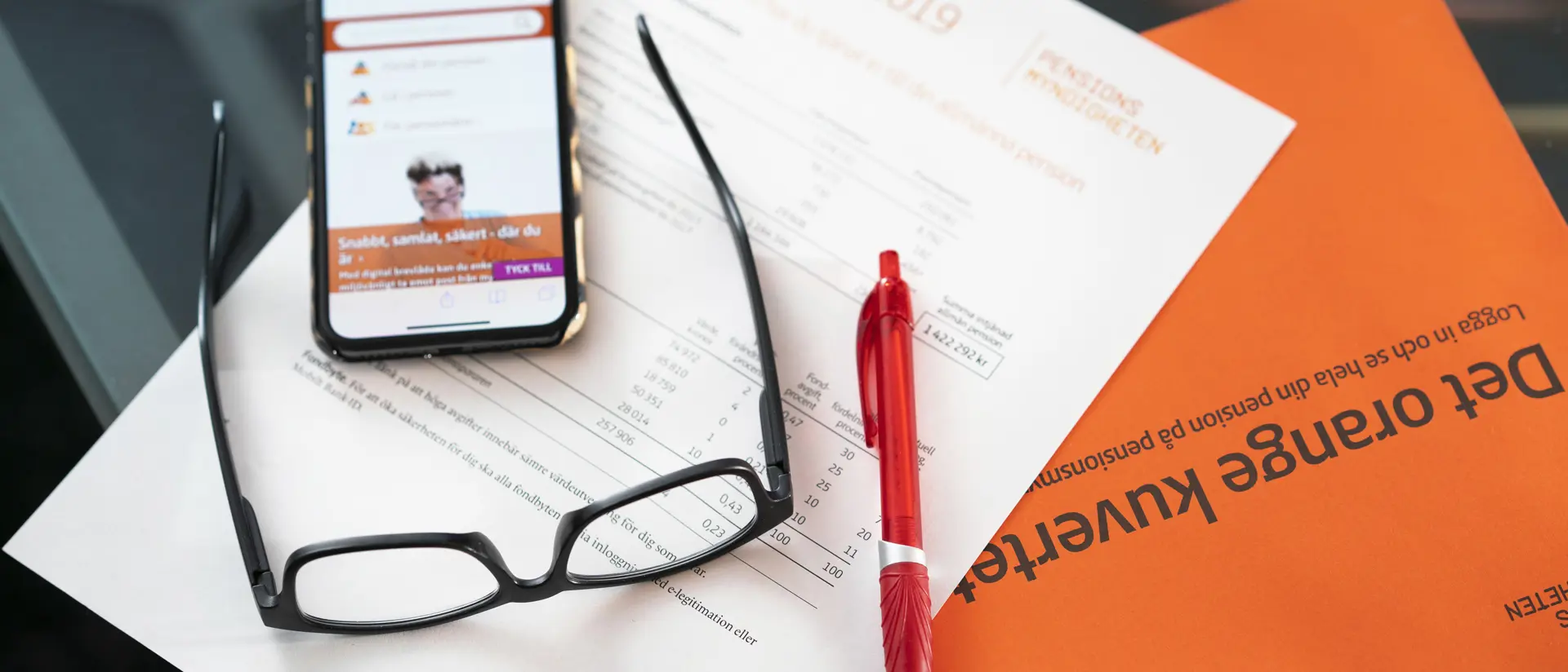 Orange kuvert, glasögon, penna och mobiltelefon.