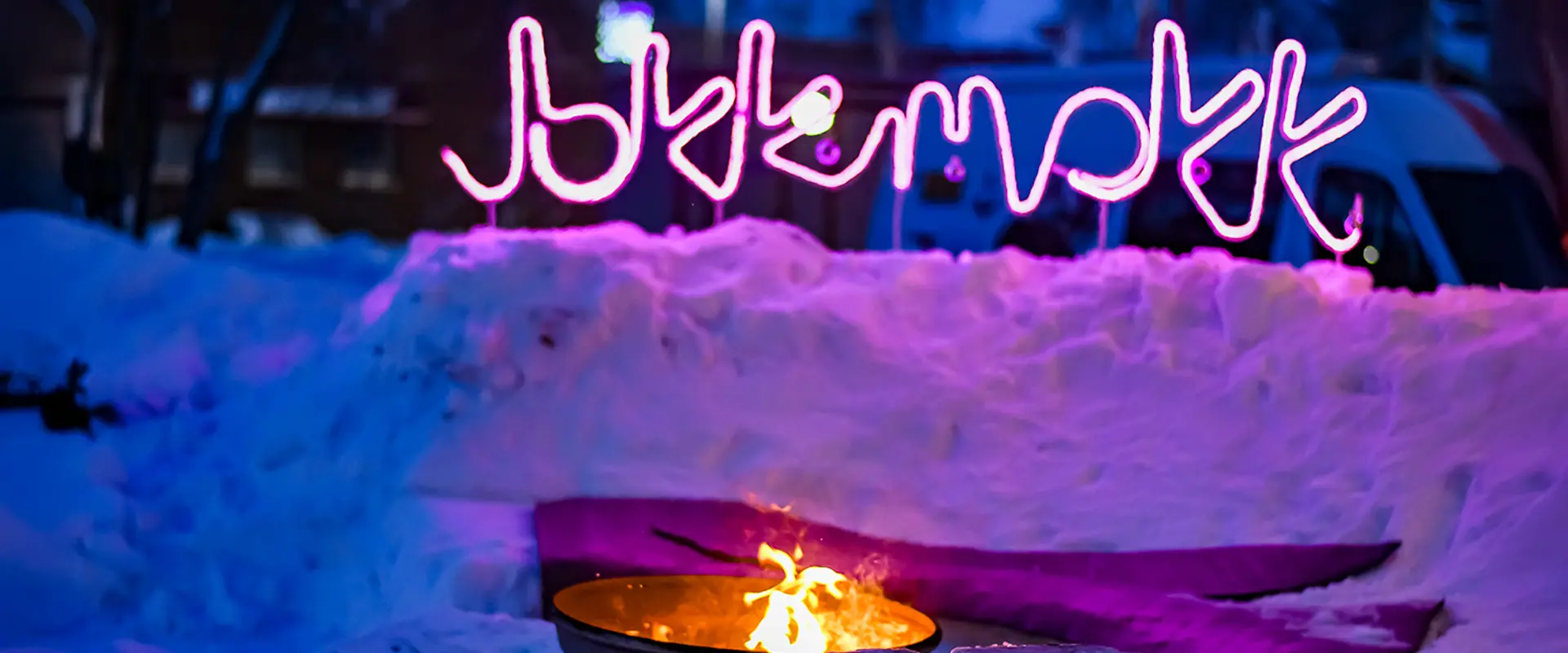 Eldplats uppbyggd av snö fotad i skymningen med en rosa neonskylt där det står