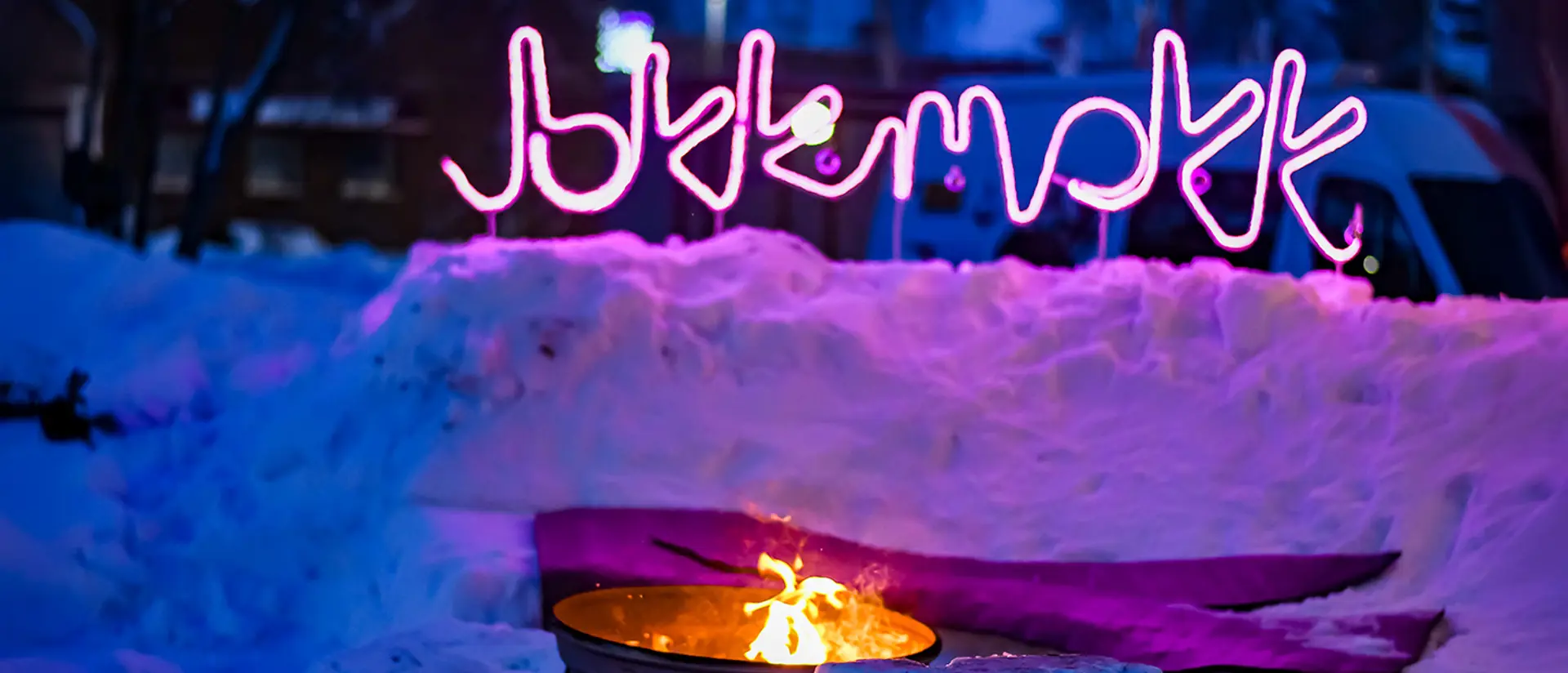 Eldplats uppbyggd av snö fotad i skymningen med en rosa neonskylt där det står